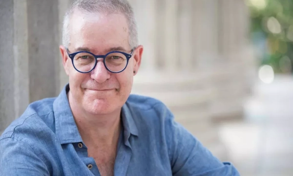A middle-aged man with glasses and short gray hair, wearing a blue button-up shirt, smiles while seated outdoors near stone columns.