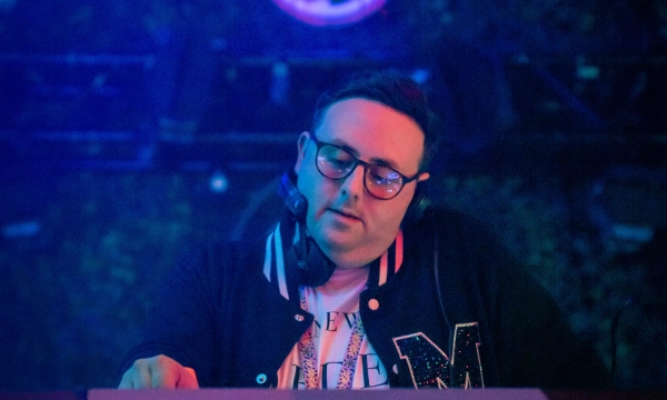 A DJ performs behind a console illuminated with colorful lights, wearing a black varsity jacket with a letter on it. A pineapple logo is displayed in the background.