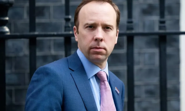 A man in a blue suit and pink tie stands outside a building with black railings, looking directly at the camera.