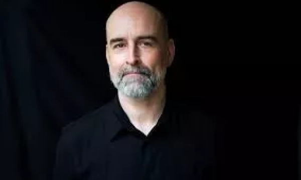 A middle-aged man with a bald head and gray beard, wearing a black shirt, stands against a dark background, looking directly at the camera.