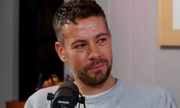 A man with short, tousled hair and a beard smiles while sitting in front of a microphone during a podcast interview.