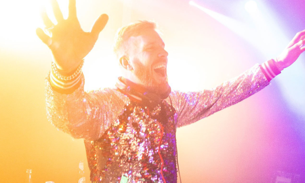 A DJ with a sparkling sequined jacket joyfully performs, reaching out to the audience amidst colorful stage lights.