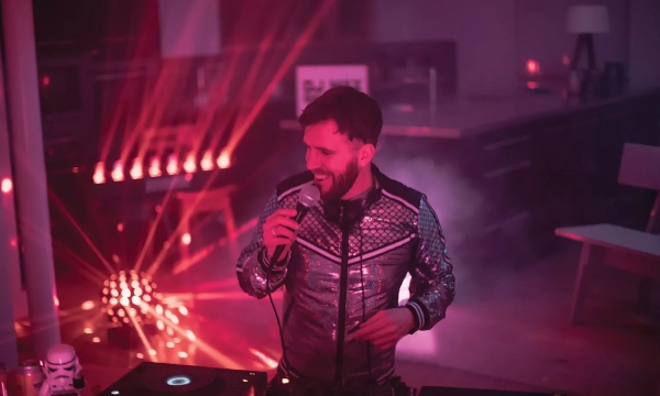 A DJ in a shiny silver jacket performs at a party, surrounded by colorful lights and smoke, creating a vibrant atmosphere.