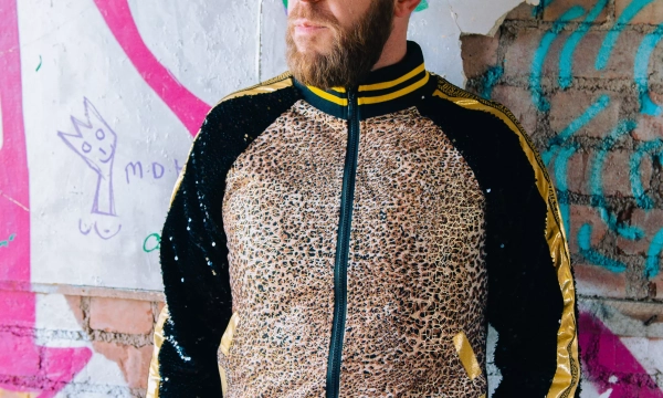 A man with a beard stands against a colorful graffiti background, wearing a leopard print jacket with black and yellow accents and blue jeans.