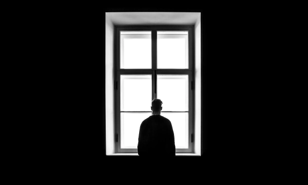 A silhouette of a person standing in front of a large window, surrounded by darkness, creating a stark contrast between light and shadow.