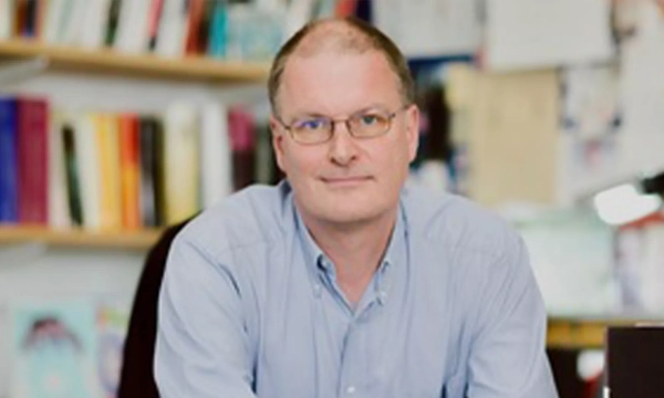 A middle-aged man with glasses and a light blue shirt seated at a desk, with bookshelves filled with various books in the background.