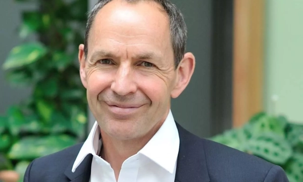 A smiling man in a suit with a white shirt, standing against a background of green plants.