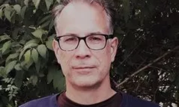 A middle-aged man with glasses, wearing a navy sweater, stands against a backdrop of leafy green plants.