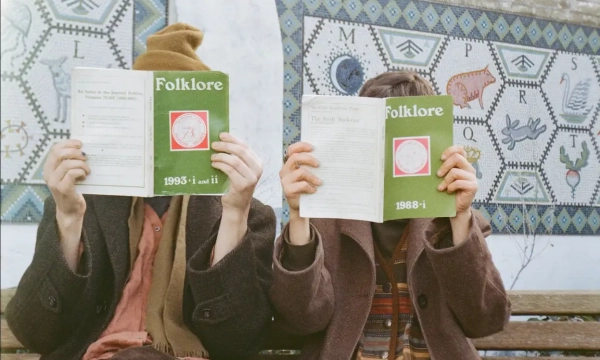 Two individuals sitting on a bench, holding up issues of a magazine titled "Folklore," with a colorful tiled mural in the background.