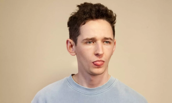 A young man with short, curly hair and a light blue sweatshirt sticks out his tongue slightly, against a neutral background.