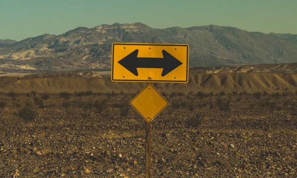A yellow directional sign with arrows pointing left and right stands in a barren landscape, with mountains in the background and rocky terrain.