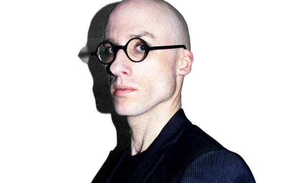 A bald man wearing round glasses and a black jacket poses against a white background, casting a distinct shadow.