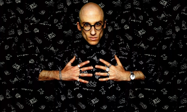 A bald man wearing round glasses and a patterned black outfit poses with his hands spread out against a matching dark background.