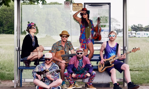 A group of six musicians dressed in colorful, vibrant outfits, posing at a bus stop with instruments, including a guitar, ukulele, and violin, against a green field backdrop.