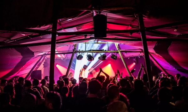 Crowd at a vibrant concert with colorful lights illuminating the stage, creating an energetic atmosphere. People are dancing and enjoying the performance.