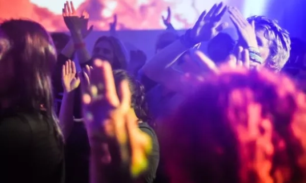 A vibrant crowd with hands raised, dancing under colorful lights at a lively music event or party.