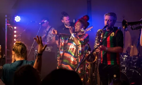 A vibrant live music performance featuring a joyful singer in a colorful outfit and musicians playing brass instruments, surrounded by an engaged audience.