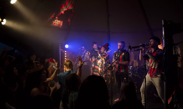 A vibrant live performance featuring a diverse band with brass instruments, colorful outfits, and a lively audience enjoying the music in a festival setting.