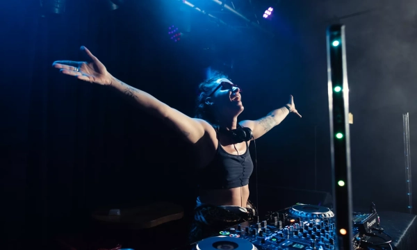 A DJ performs enthusiastically at a nightclub, with arms outstretched, surrounded by lighting and equipment, creating an energetic atmosphere.