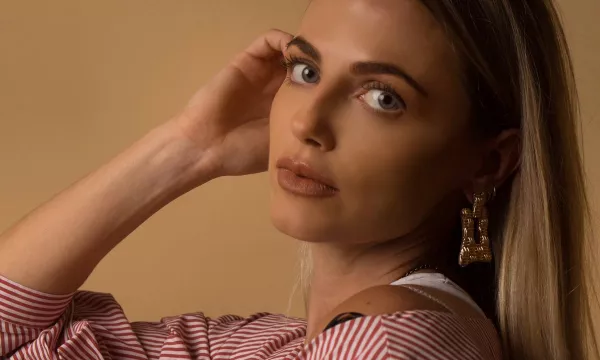 A close-up portrait of a woman with long hair, wearing a striped blouse and large earrings, gazing thoughtfully at the camera against a neutral background.