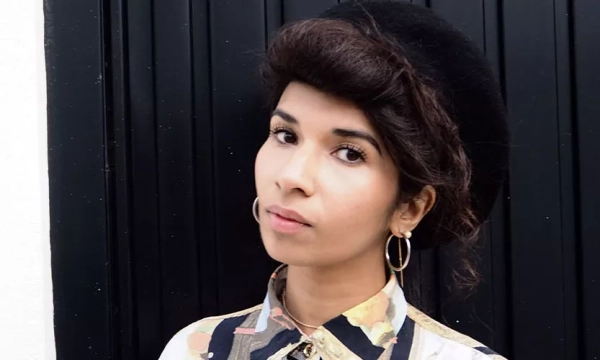 A woman with dark, curly hair styled in a vintage fashion, wearing a patterned blouse and large hoop earrings, stands in front of a black door.
