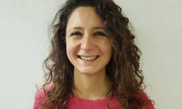Smiling woman with curly hair wearing a pink sweater, standing against a plain background.