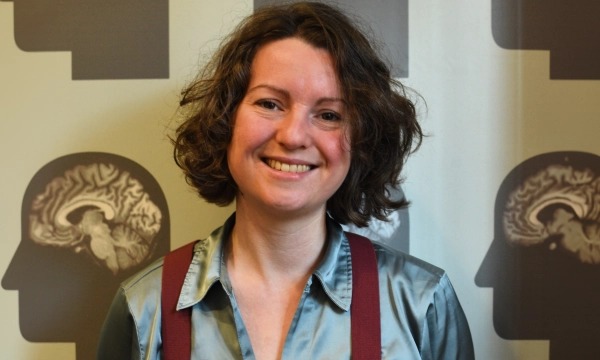A smiling woman with curly hair wears a light blue shirt and red suspenders, standing in front of a backdrop featuring brain illustrations.