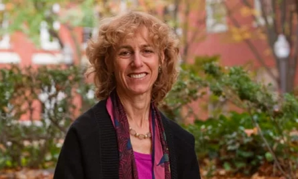 A smiling woman with curly hair, wearing a black cardigan and scarf, stands outdoors in a leafy setting, showcasing a warm and friendly demeanor.