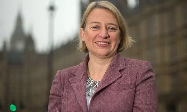 A woman with short blonde hair smiles confidently while standing in front of a historic building, wearing a pink blazer and necklace.