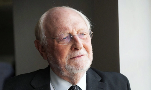 An elderly man with glasses and a beard, wearing a suit, gazes thoughtfully out a window.