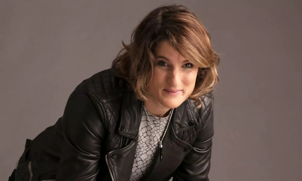Portrait of a smiling woman with shoulder-length wavy hair, wearing a leather jacket, leaning slightly forward against a neutral backdrop.