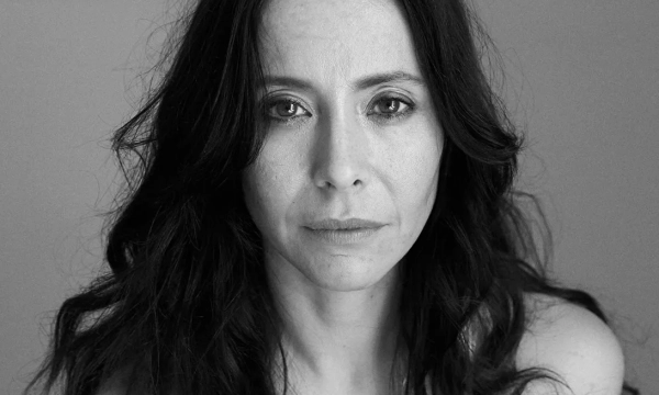 A woman with long dark hair gazes intently at the camera, wearing a simple necklace that reads "StarLuck." The background is minimalist and softly lit.