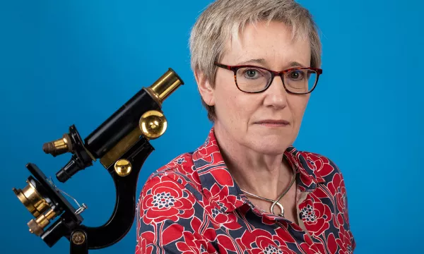 A woman with short grey hair and glasses poses in front of a blue background, next to a vintage microscope, wearing a floral-patterned shirt.