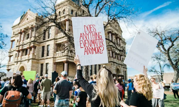 A crowd gathers in front of a historic building, with two individuals holding signs, one reading "Does anything even matter anymore?" under a clear blue sky.