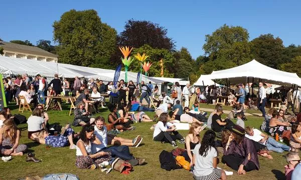 A lively outdoor festival scene with numerous attendees relaxing on the grass, surrounded by tents and colorful decorations, under a clear blue sky.