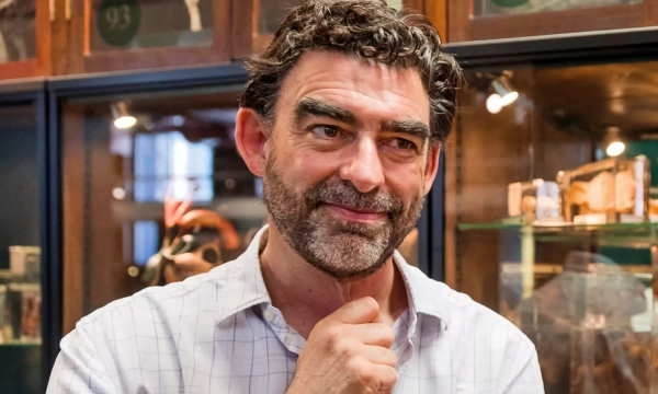 A smiling middle-aged man with curly hair and a beard, dressed in a white shirt, stands thoughtfully in a museum setting with display cases in the background.