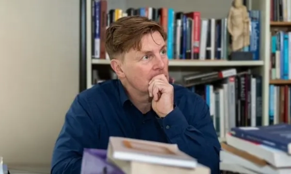 A thoughtful man in a blue shirt sits at a table surrounded by stacks of books, looking off into the distance with a contemplative expression.