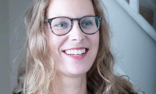 A smiling woman with long, wavy hair and glasses, wearing a patterned blouse, stands in a bright interior with a staircase in the background.