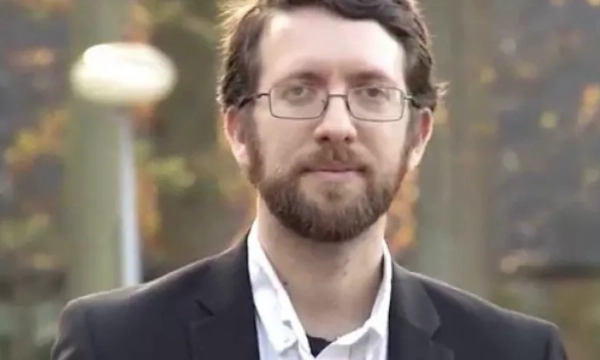 A man with glasses and a beard, wearing a black suit jacket, stands outdoors with a blurred background of autumn leaves.