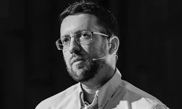 A thoughtful man with glasses and a microphone clip, engaged in a serious discussion or presentation. The background is dark, emphasizing his expression.