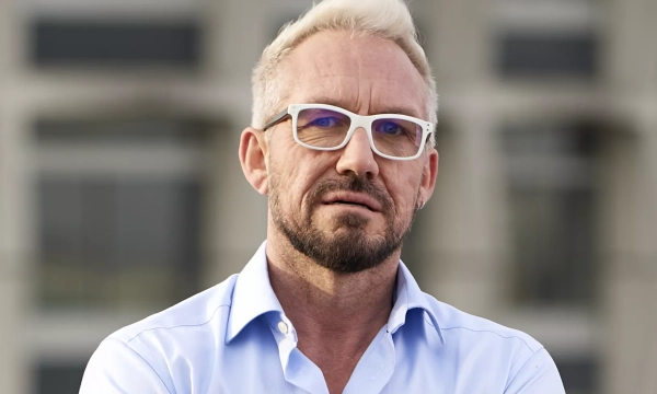 A man with short platinum blonde hair and glasses wearing a light blue button-up shirt, standing outdoors against a blurred urban background.