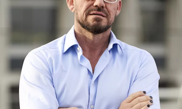 A man stands confidently with his arms crossed, wearing a light blue shirt and white glasses, with a watch on his wrist and nail polish on his fingers.