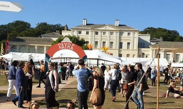 A bustling outdoor festival scene with people enjoying activities, tents, and directional signs, set against an elegant historic building in the background.