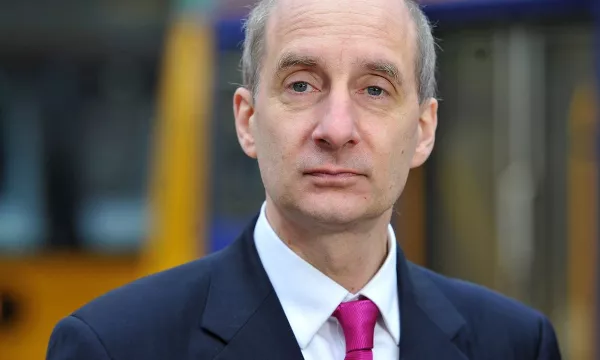 A man in a dark suit and pink tie stands outdoors, looking seriously at the camera, with a blurred background of vehicles.