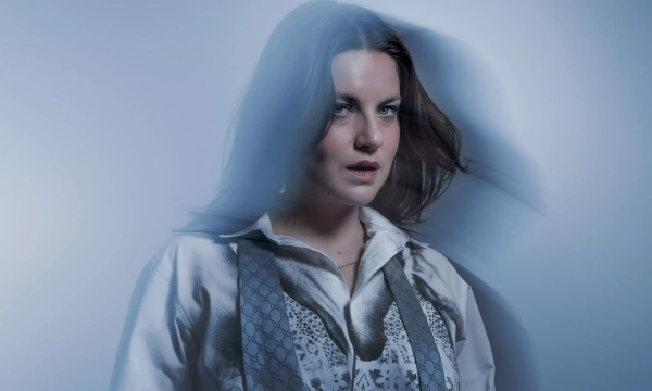 A woman with long hair stands against a soft blue background, wearing a patterned shirt and a light gray scarf, with a contemplative expression.