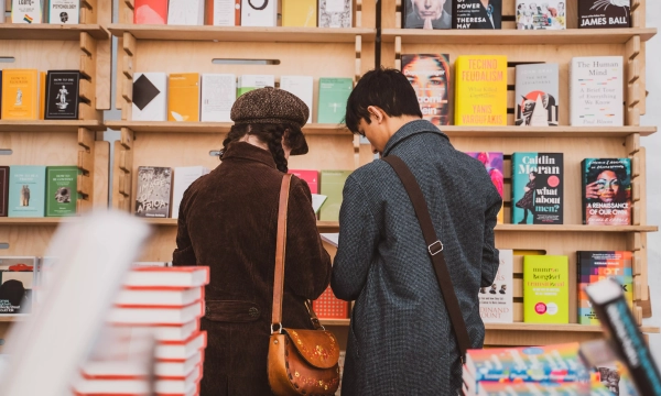 Two people browsing books in a cozy bookstore, with a variety of colorful book covers displayed prominently on the shelves behind them.