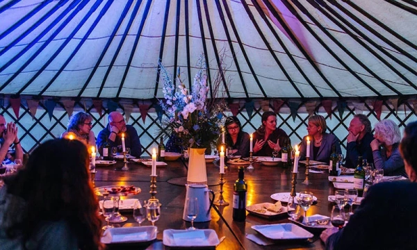 A dinner party inside a yurt, featuring guests seated around a large table adorned with candles, flowers, and wine, illuminated by soft colored lights.