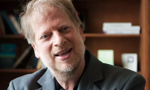 A man with light hair and a beard, wearing a dark suit, smiles while speaking in an office with bookshelves in the background.