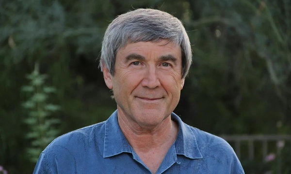 A smiling man with gray hair, wearing a blue shirt, stands outdoors with greenery in the background.