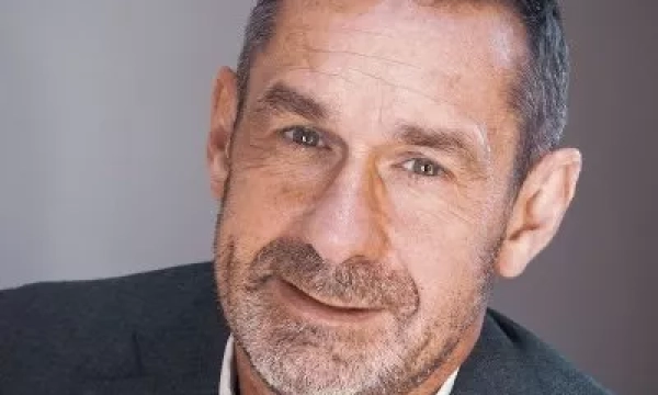 A middle-aged man with a short beard and grey hair, wearing a grey suit, smiling against a neutral background.